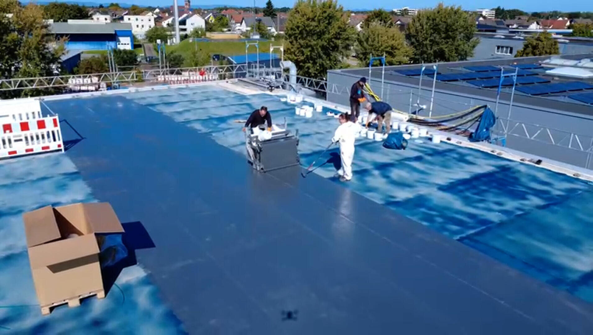 Drei Arbeiter sind dabei, ein großes schwarzes Material auf einem Dach zu installieren oder zu verlegen, das von Gebäuden und Bäumen umgeben ist und auf dem Geräte und Kisten verstreut sind. Der Himmel ist klar und sonnig.