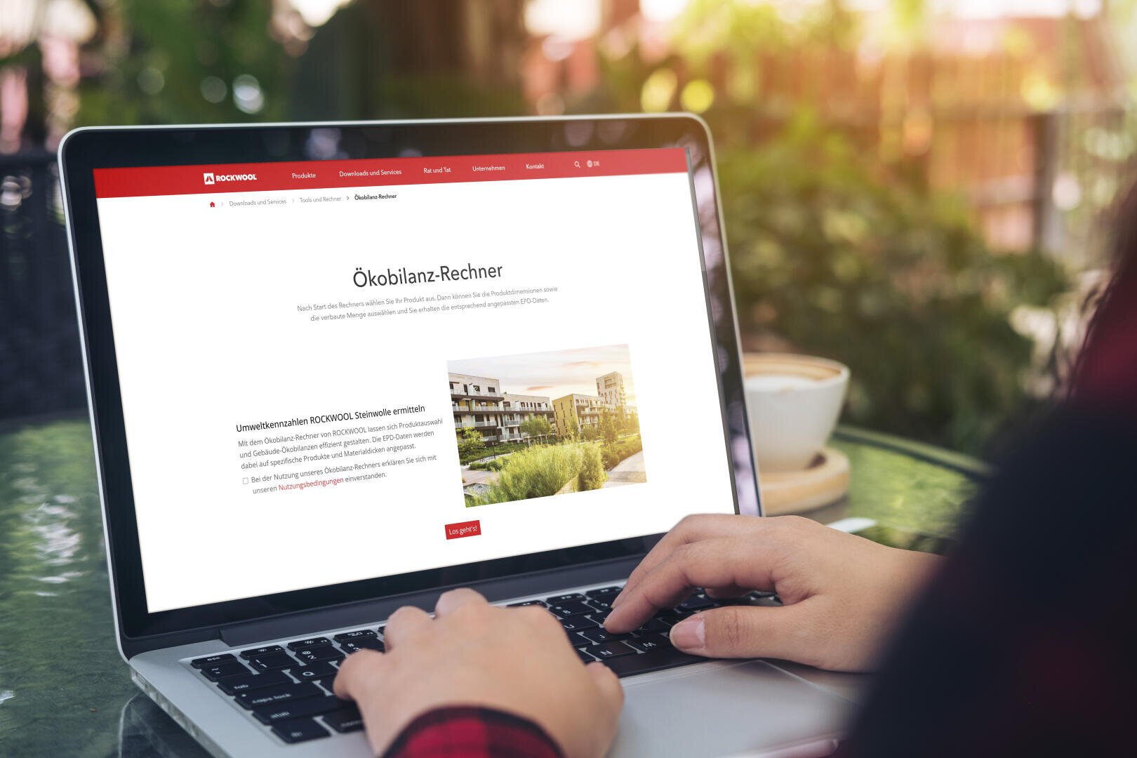 A person types on a laptop displaying a German website with the title Ökobilanz-Rechner and a picture of modern buildings; a coffee cup and green plants can be seen in the background.