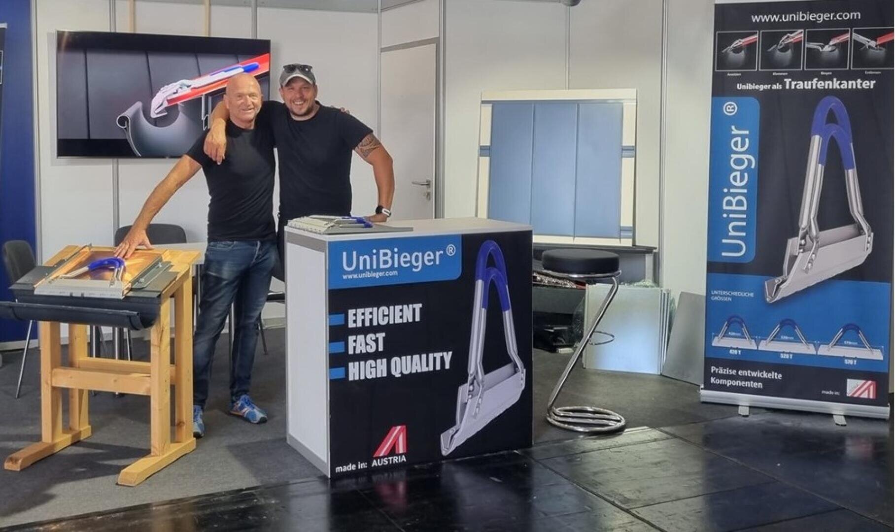 Two smiling men stand together at an exhibition stand displaying UniBieger metal bending tools. Marketing banners and product samples can be seen in the background, emphasizing efficiency, speed and quality.