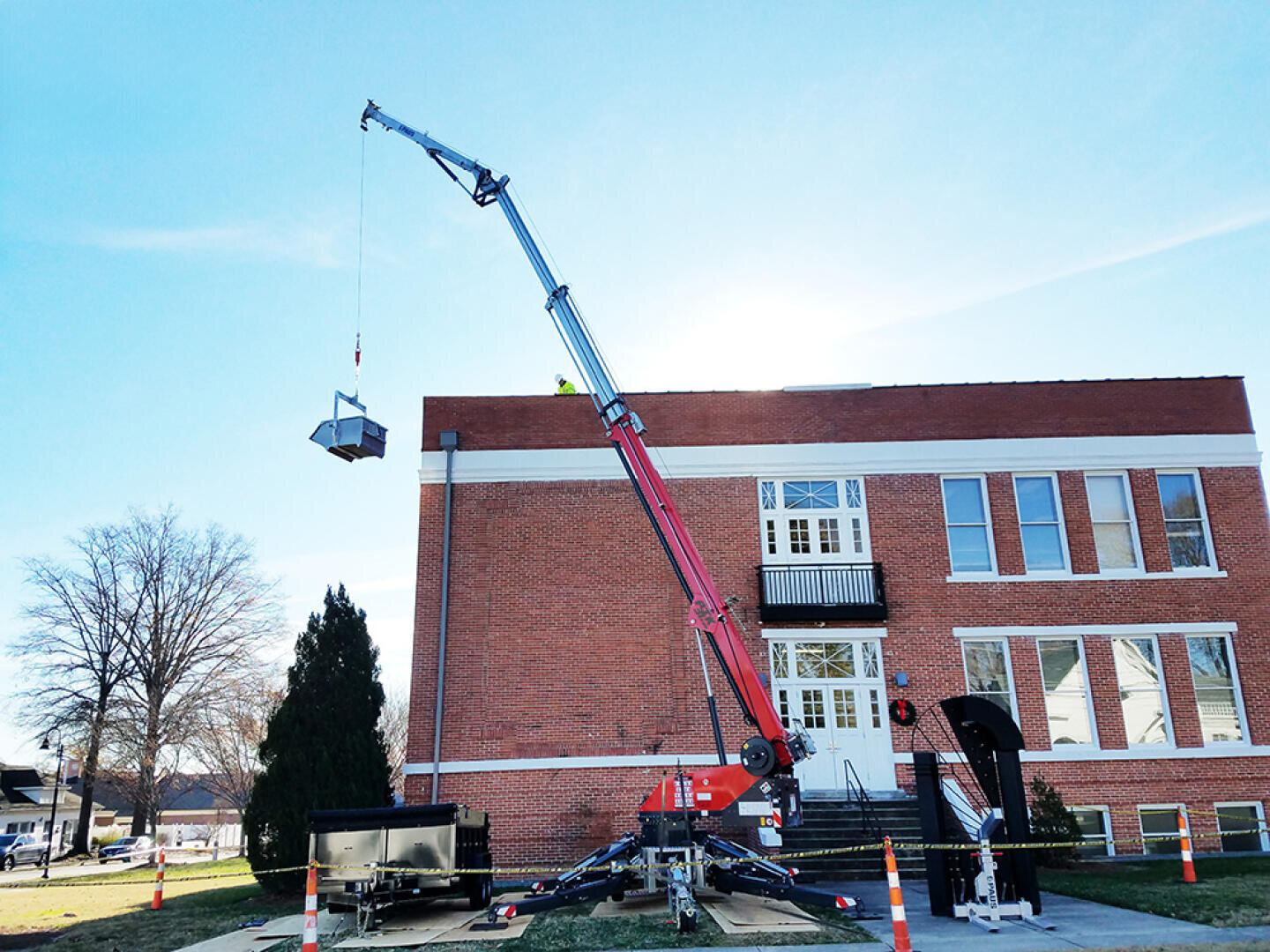 Ein roter Kran hebt Geräte auf das Dach eines Backsteingebäudes. Der Kran ist auf dem Rasen mit Auslegern aufgestellt, und orangefarbene Sicherheitskegel markieren den Bereich. Ein blattloser Baum steht in der Nähe unter einem klaren Himmel.