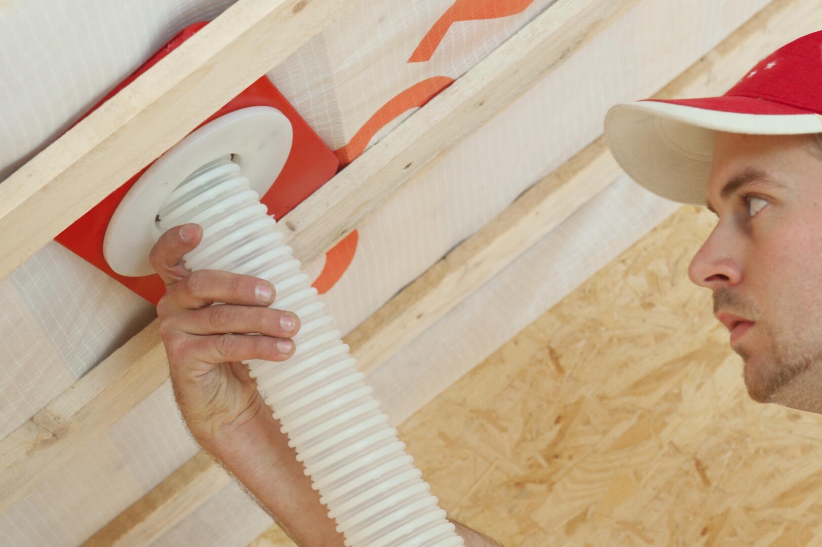A man with a red and white cap installs a flexible white ventilation pipe through a ceiling construction made of wooden beams and chipboard. With one hand, he secures the pipe in a red mounting plate.