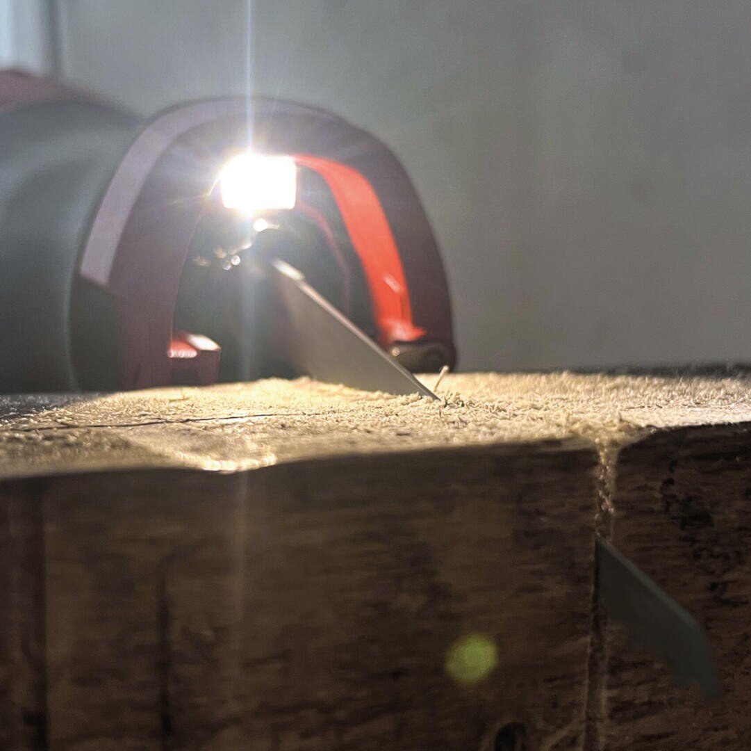 Nahaufnahme einer handgeführten elektrischen Säge, die ein Stück Holz durchschneidet, wobei ein helles Licht aus dem Werkzeug leuchtet, das Sägespäne und Holzmaserung beleuchtet.