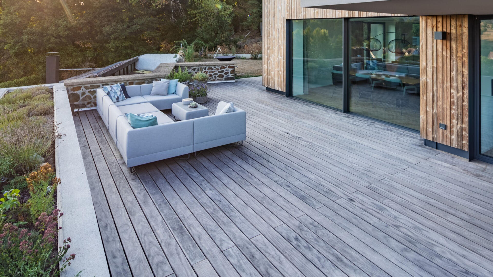 Eine moderne Holzterrasse mit einem hellgrauen Outdoor-Sofa, Kissen und einem Couchtisch, neben einem Haus mit großen Glastüren. Die Terrasse blickt auf Grünflächen und einen angelegten Gartenbereich.