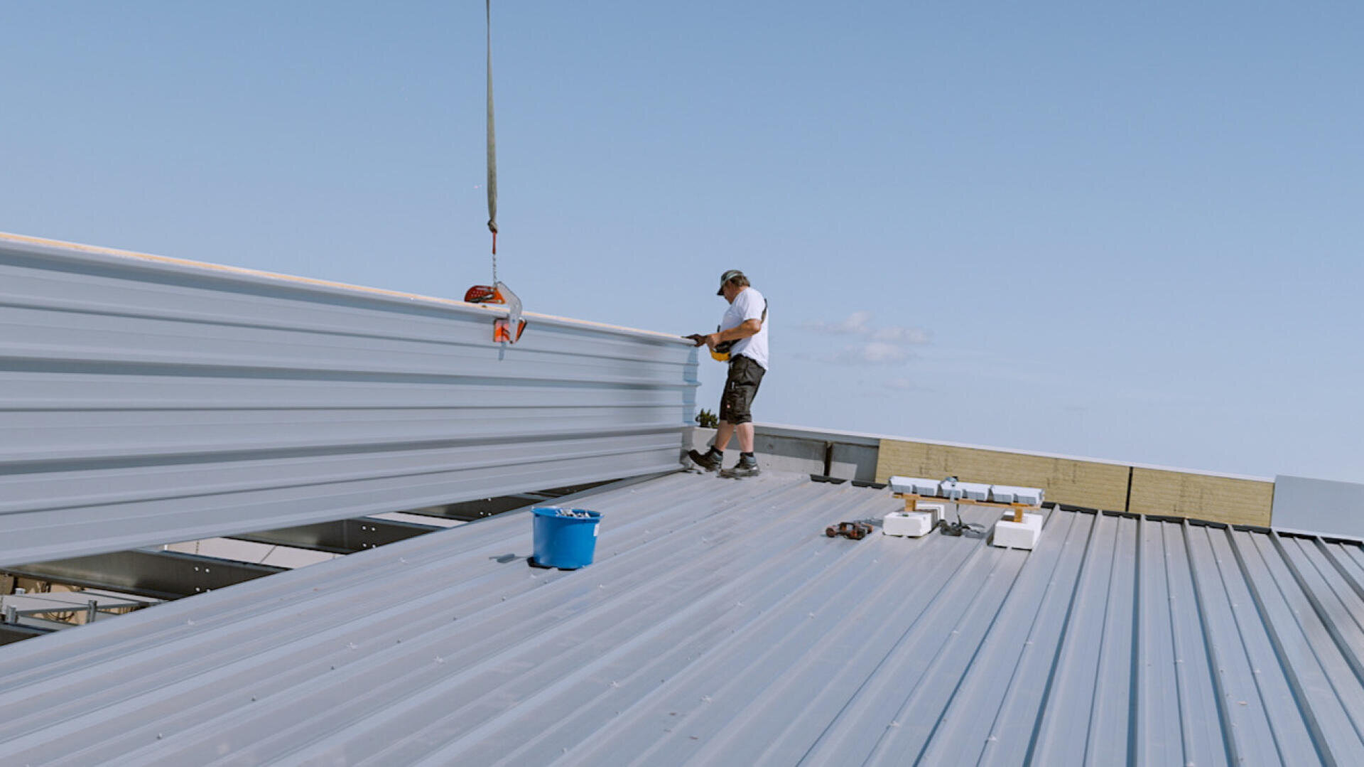 A worker in shorts and a cap stands on a metal roof and assembles large metal plates with a crane under a blue sky. There are tools and a blue bucket on the roof. SO THAT EVERYTHING FITS. The lettering ROMA appears in the corner.