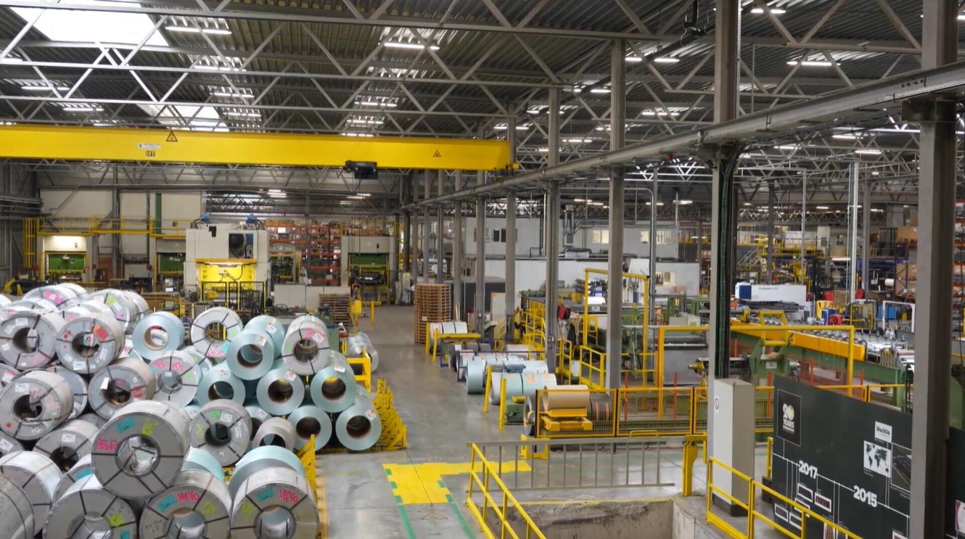 Ein geräumiges Industrielager mit Stapeln von gewalzten Blechen, gelben Sicherheitsgeländern, Brückenkränen und organisierten Arbeitsplätzen unter einer hohen Stahlträgerdecke.