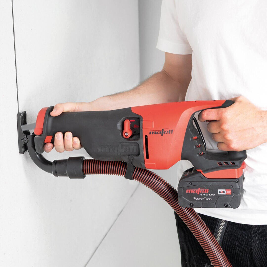 Eine Person benutzt ein rot-schwarzes Elektrowerkzeug mit einem Schlauchaufsatz, um in eine weiße Wand zu schneiden. Das Gerät ist mit Mafell beschriftet und hat einen Akku und einen Schlauch für die Staubabsaugung.