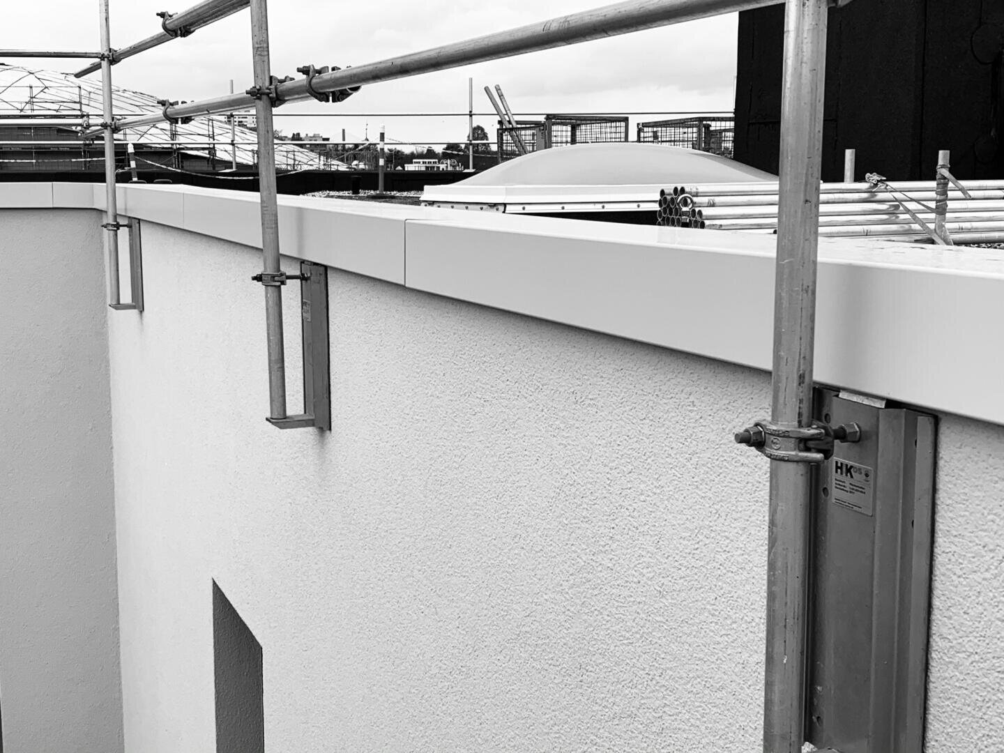 A black and white photo shows the corner of a building with scaffolding set up along the top edge. The wall has a smooth, textured surface and there is a flat roof with construction materials visible.