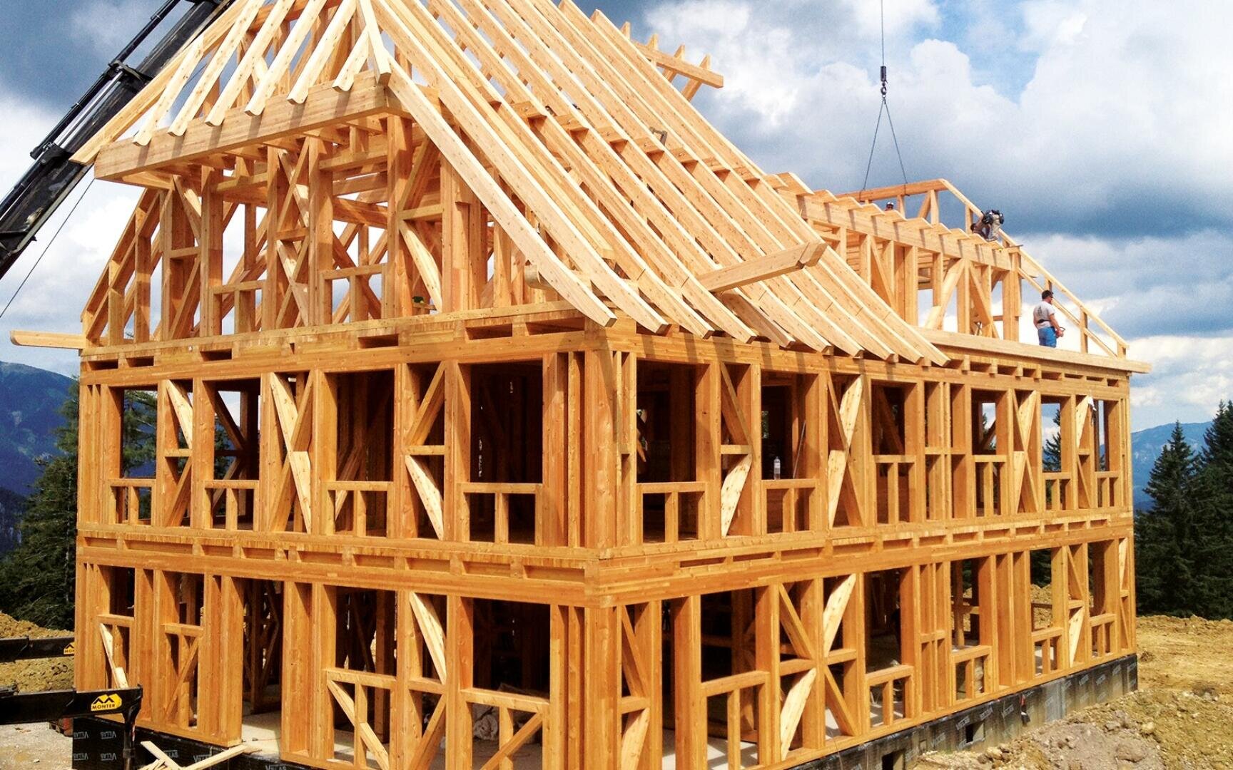 A two-story wooden house under construction with exposed timber frame and roof structure, with workers on the upper floor and a crane for support. The building is set against a background of trees and a cloudy sky.