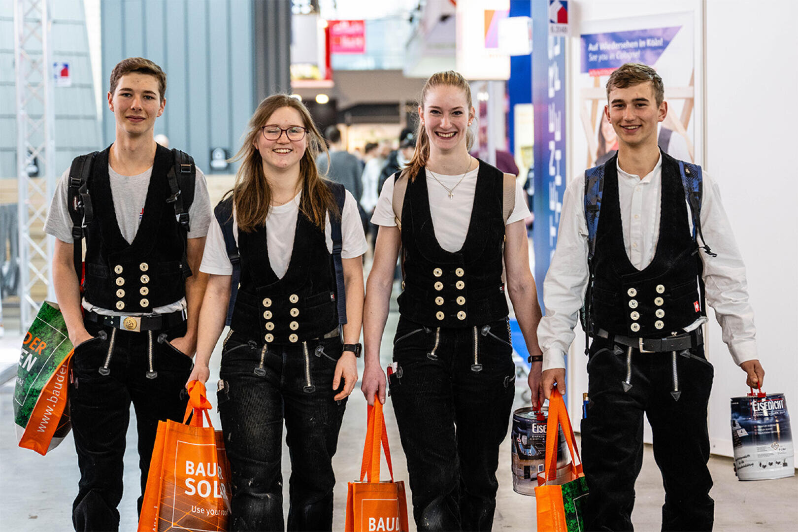 Four young people wearing matching traditional black vests, white shirts and dark pants walk into the interior smiling and carrying bright orange carrier bags. They appear to be at an event or exhibition.
