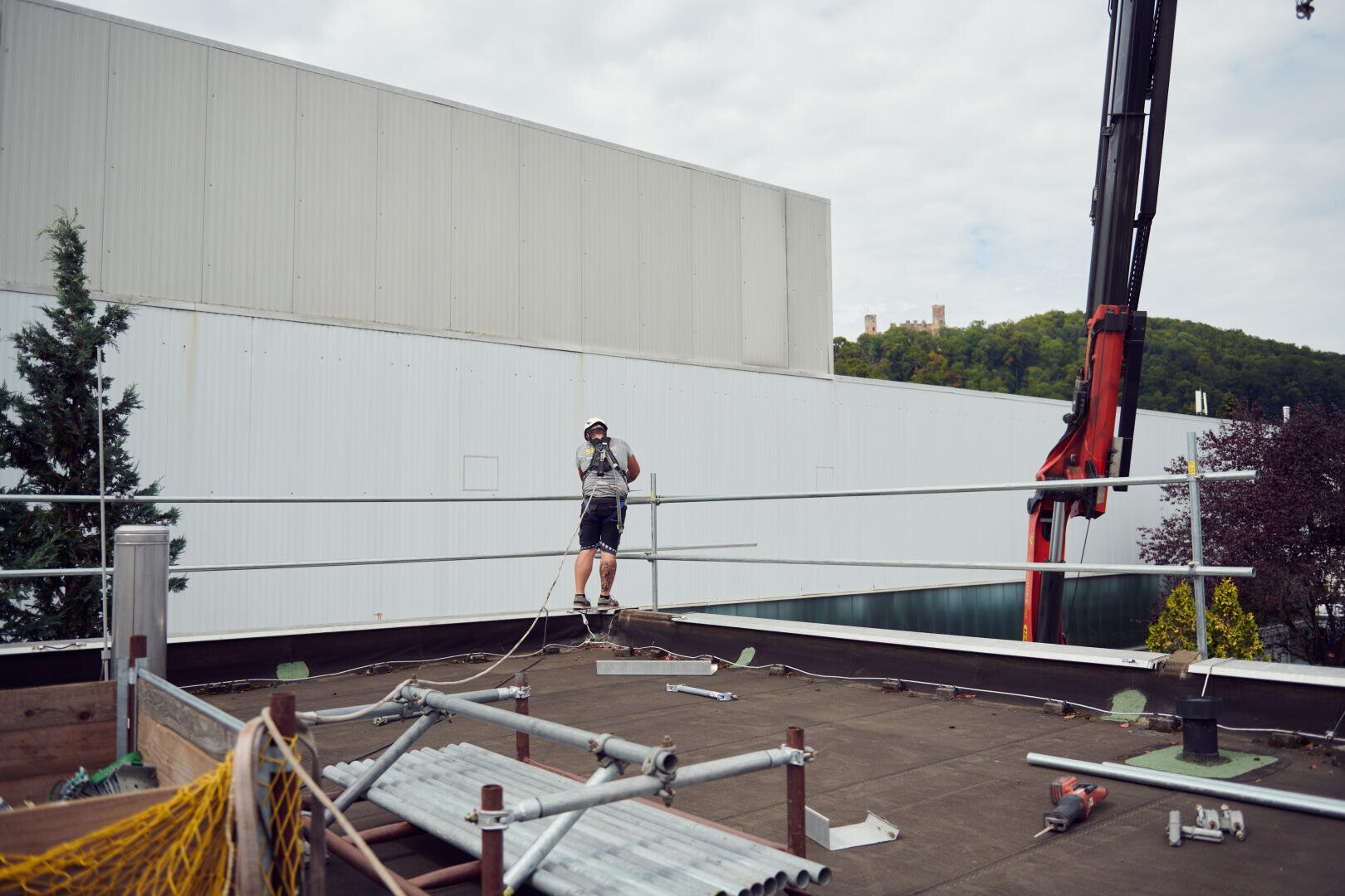 Ein Bauarbeiter in Schutzkleidung steht auf einem Dach und hält sich an Kabeln fest, umgeben von Baumaterialien und Werkzeugen. In der Nähe ragt ein Kranarm in die Höhe, im Hintergrund sind Bäume und Industriegebäude zu sehen.