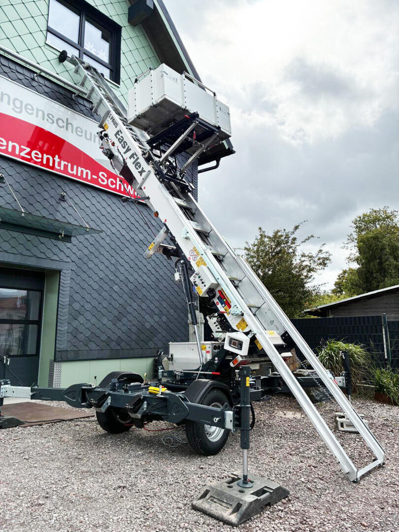 Eine mobile Hebebühne mit einer ausziehbaren Leiter und einem großen Eimer steht vor einem Gebäude auf Kies. Die Struktur wird von stabilisierenden Beinen gestützt, und der Himmel ist bedeckt.