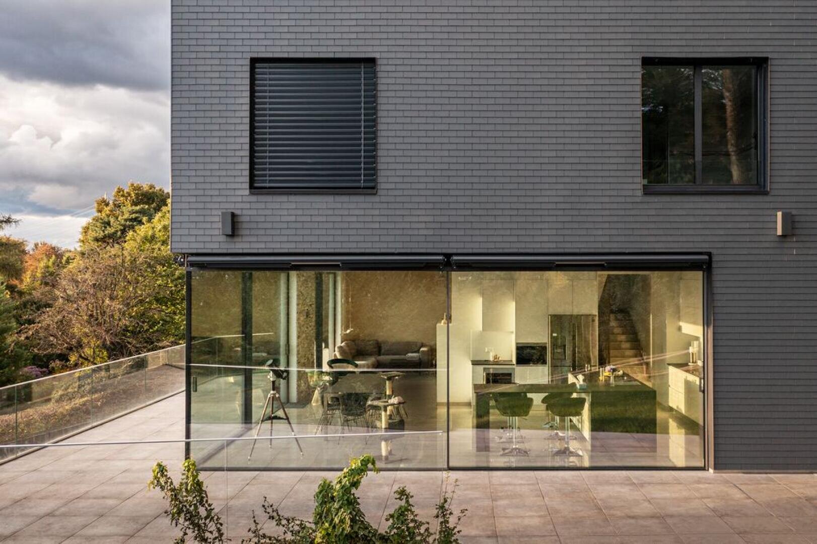 A modern, gray brick house with large glass windows provides a view of a stylish kitchen and living area inside. The spacious outdoor terrace is bordered by a glass railing and trees can be seen in the background.
