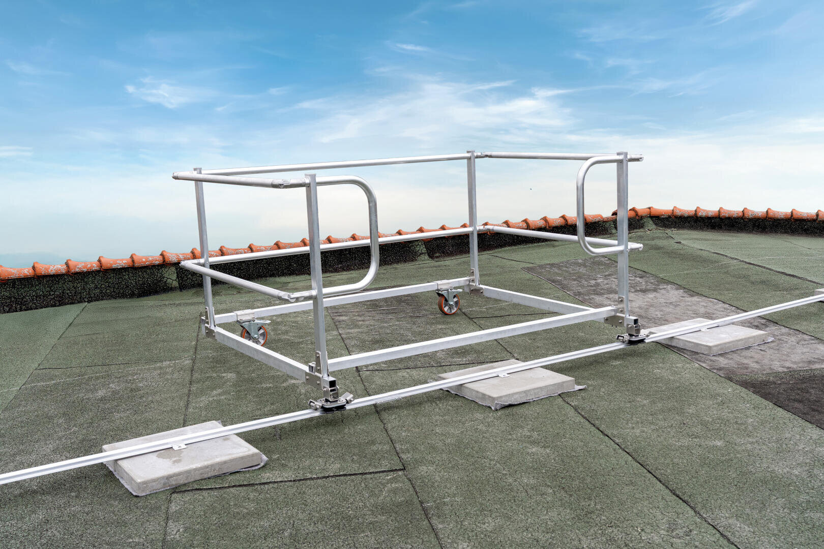 Auf einem Flachdach mit grünem Abdichtungsmaterial wird unter blauem Himmel ein Sicherheitsgeländersystem aus Metall installiert, das mit Rädern versehen und mit beschwerten Sockeln gesichert ist.