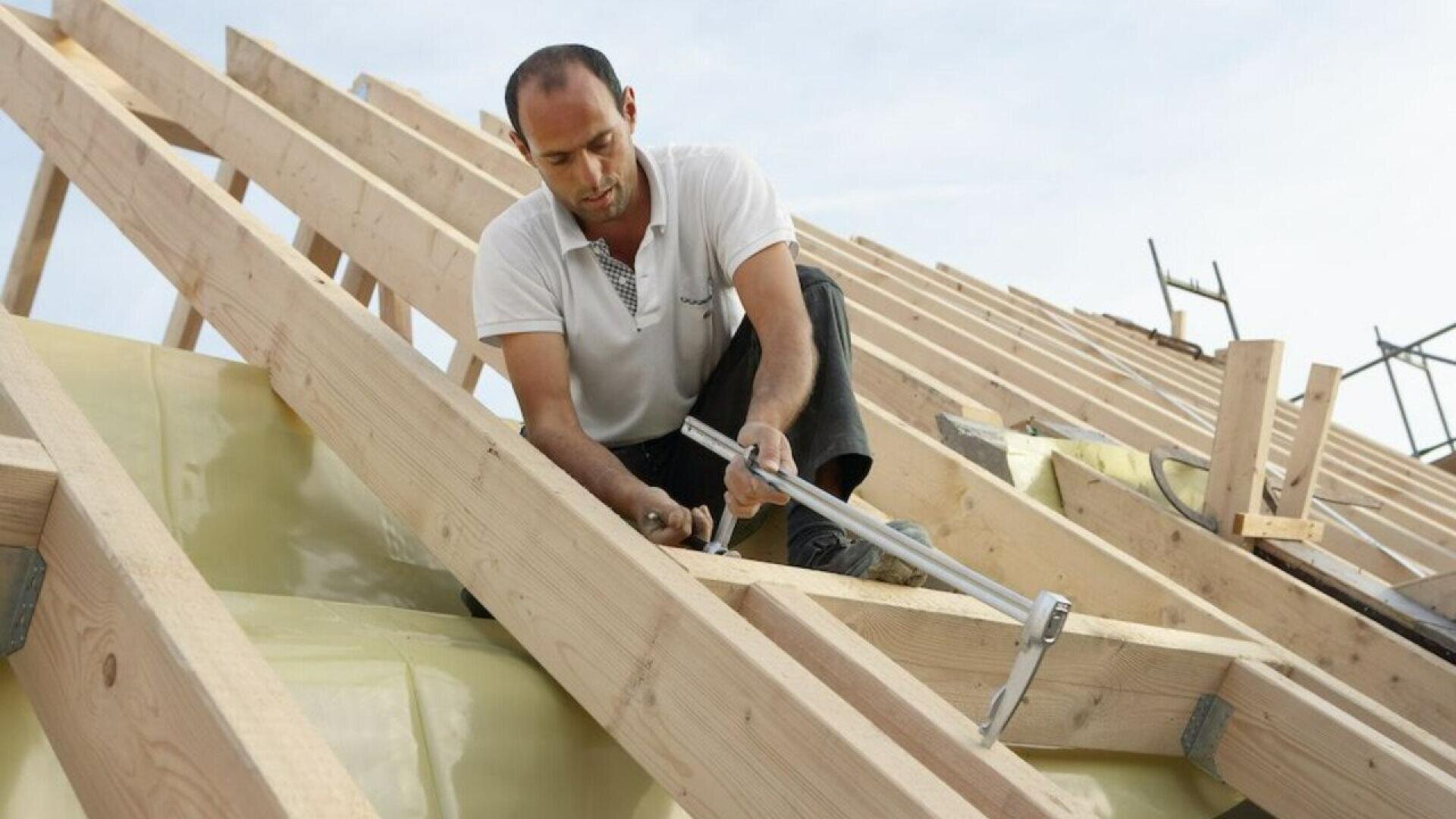 A man in a white shirt uses a hammer to work on the wooden frame of a roof under construction, with building materials and blue sky in the background.