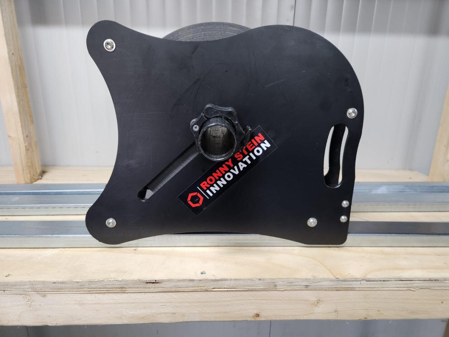 Ein schwarzer Spulenhalter mit der Aufschrift RONNY STEIN INNOVATION steht auf einem Holzregal, in dem eine große Spule mit Material teilweise sichtbar ist. Der Halter hat eine Griffaussparung und steht an einer gewellten Wand.
