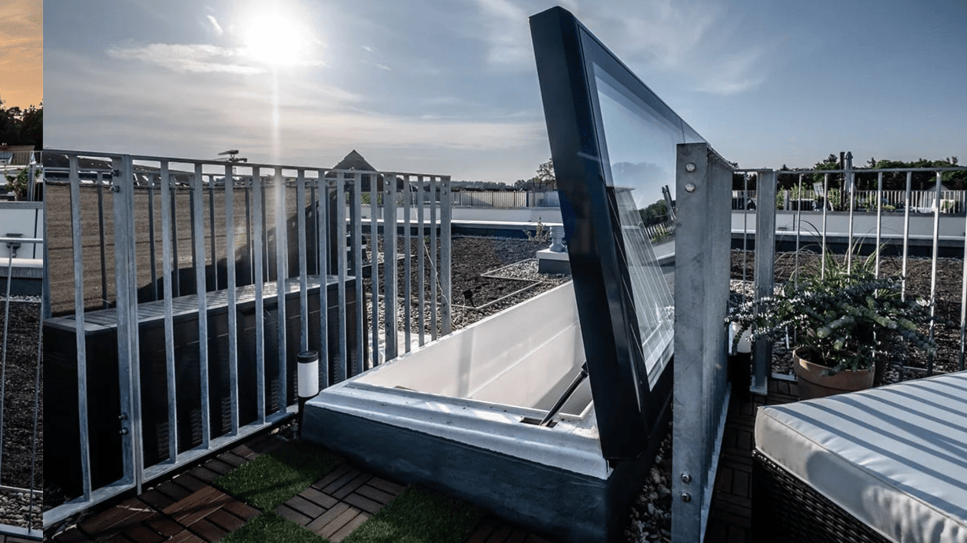 Ein modernes Dach mit einer teilweise geöffneten Glasluke, Metallgeländern, Topfpflanzen und Sonnenlicht im Hintergrund.