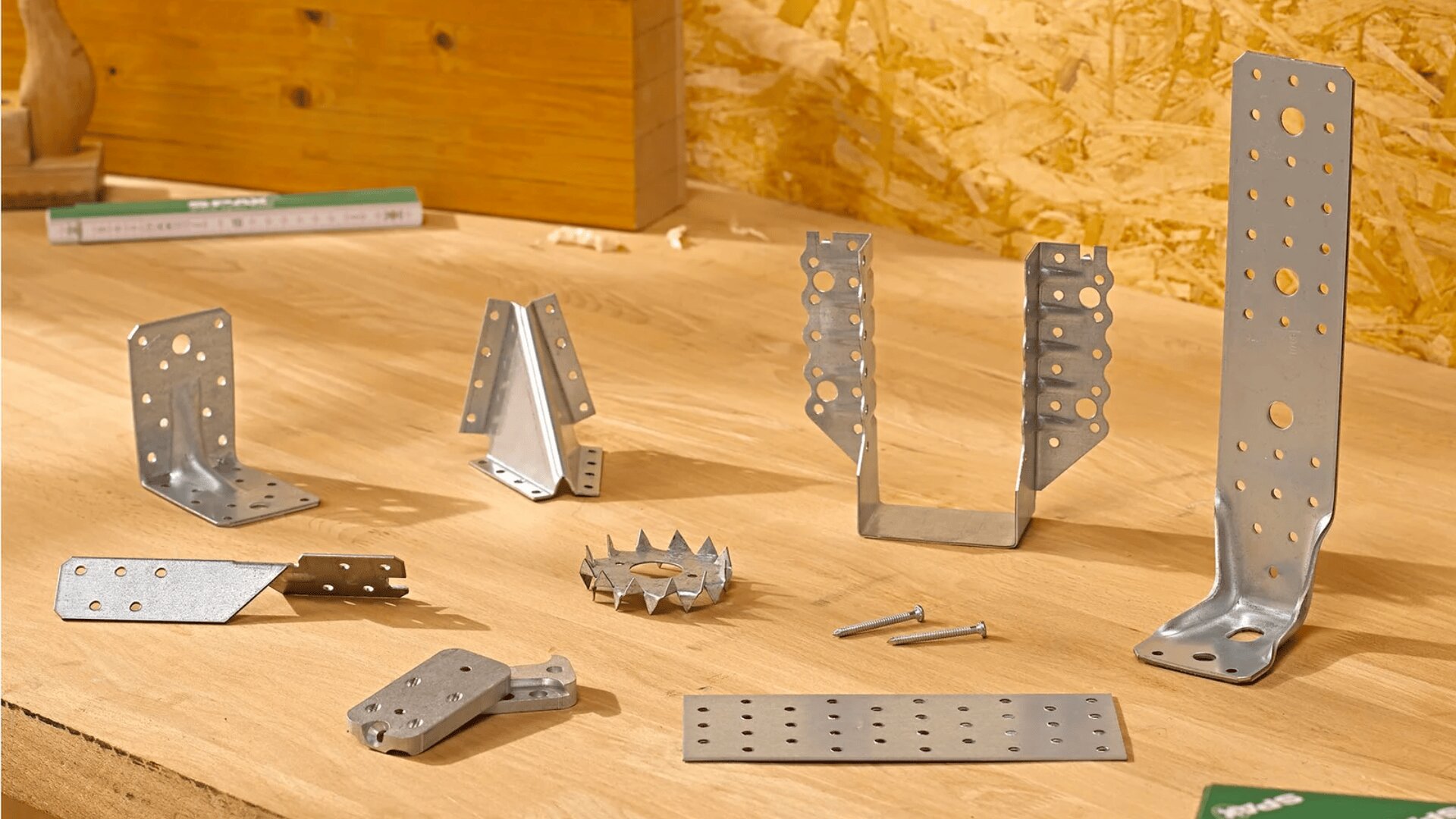 Verschiedene Metallkonstruktionswinkel, Verbindungselemente und Befestigungselemente sind auf einer Holzwerkbank angeordnet. Auch einige Schrauben und Nägel sind sichtbar. Im Hintergrund sind eine holzgetäfelte Wand und weitere Werkstattgegenstände zu sehen.
