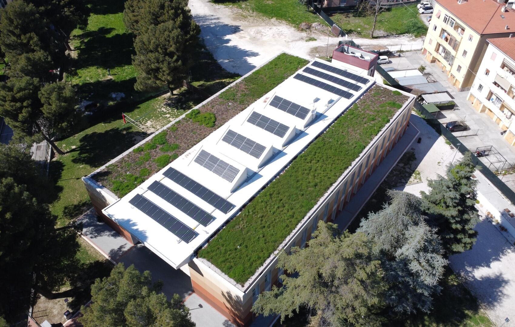 Luftaufnahme eines rechteckigen Gebäudes mit einem begrünten Dach und mehreren Sonnenkollektoren, umgeben von Bäumen, Grünflächen und nahe gelegenen Wohngebäuden.