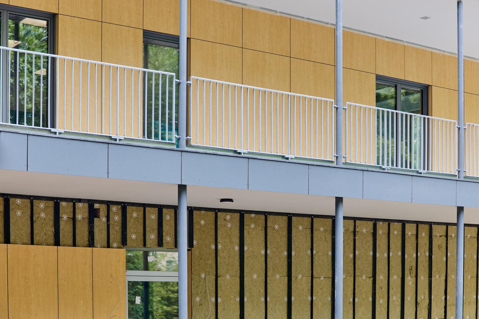 Modern building façade under construction with yellow insulation panels partially installed in the lower section and finished timber cladding with white railings and large windows in the upper section.