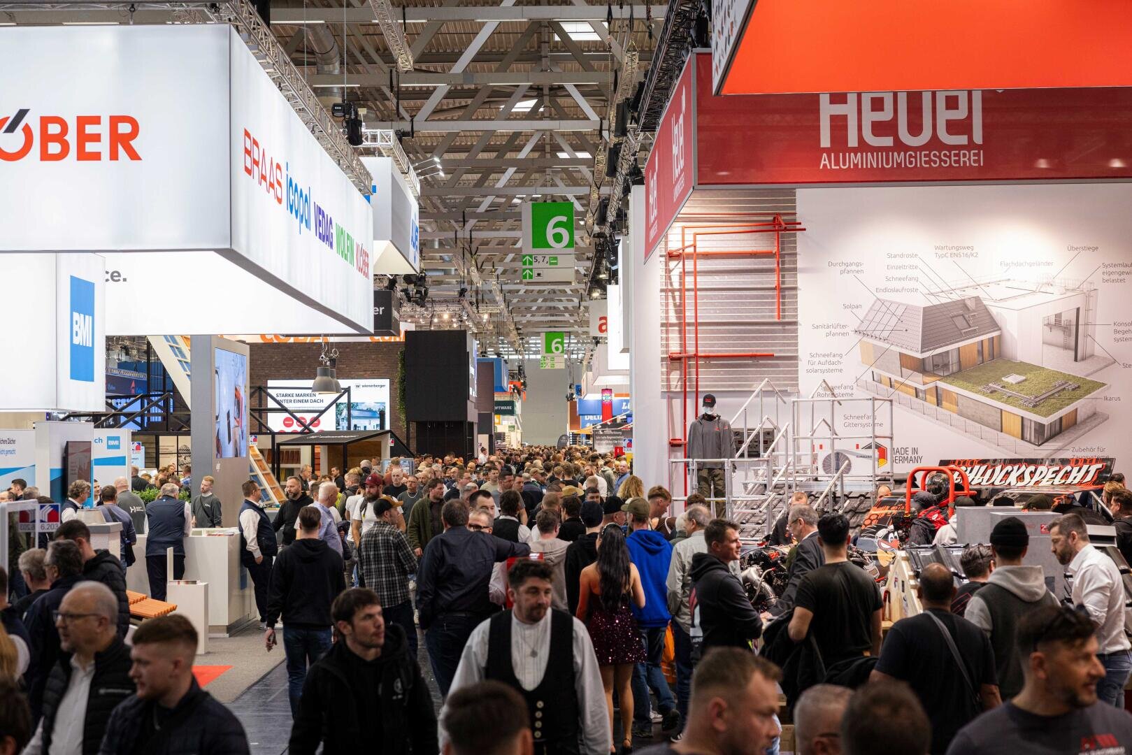 Eine große Menschenmenge läuft durch eine belebte Messe- oder Kongresshalle, in der verschiedene Firmenstände, Banner und Bauauslagen zu sehen sind.