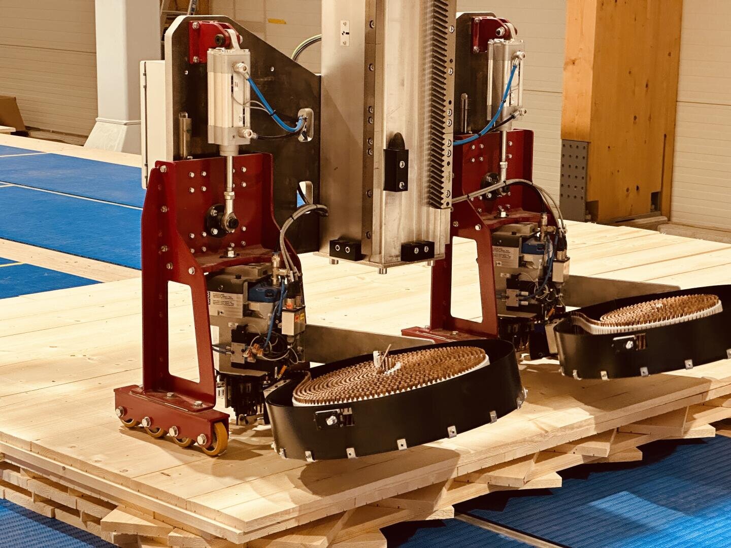 Nahaufnahme eines Roboters mit roten Metallteilen und Kabeln, der auf gestapelten Holzbrettern positioniert ist und möglicherweise im Baugewerbe oder bei der automatischen Holzverarbeitung eingesetzt wird. Blaue Matten bedecken den Boden im Hintergrund.