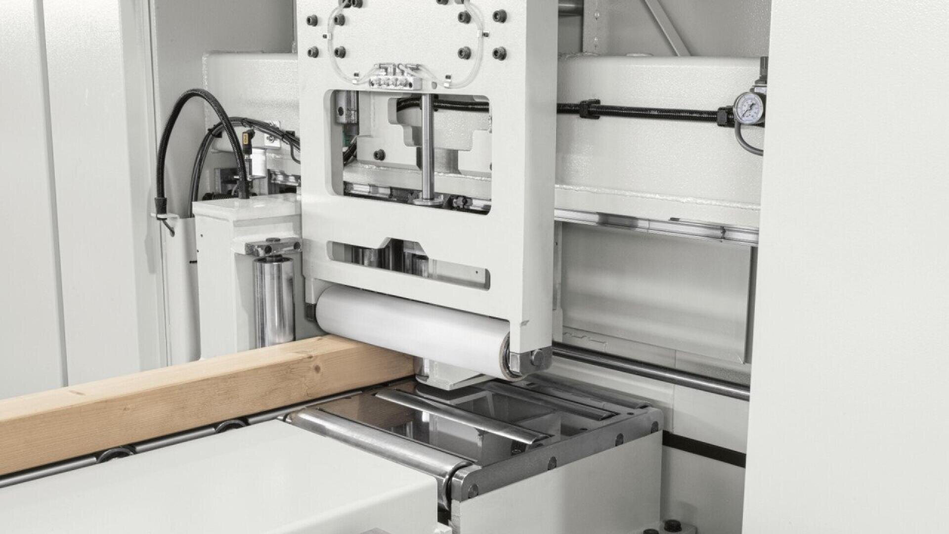 A close-up view of an industrial woodworking machine processing a wooden beam, with metal components and rollers visible in a clean, white setting.