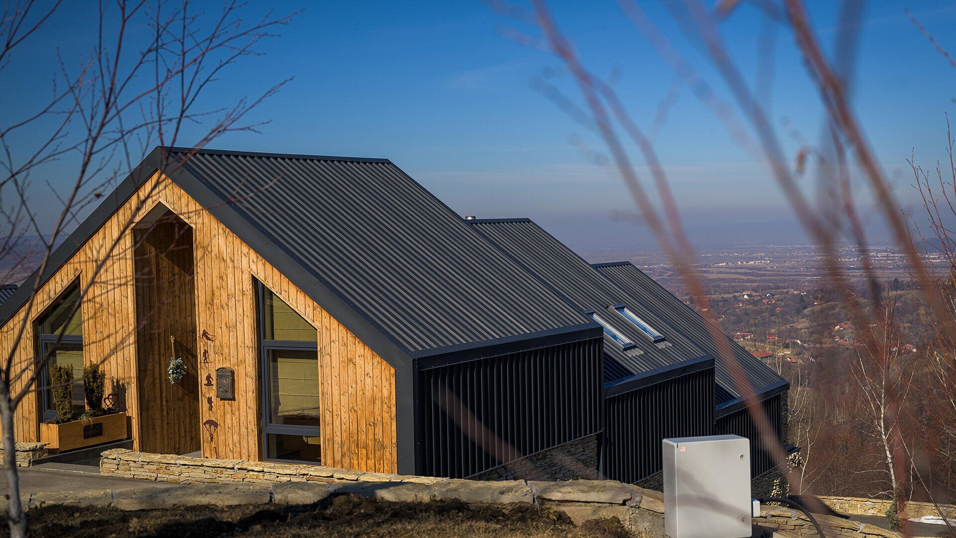 Eine moderne Holzhütte mit schwarzem Metalldach steht auf einem Hügel mit Blick auf ein Tal und eine entfernte Stadt unter einem klaren blauen Himmel, mit kahlen Baumzweigen im Vordergrund.