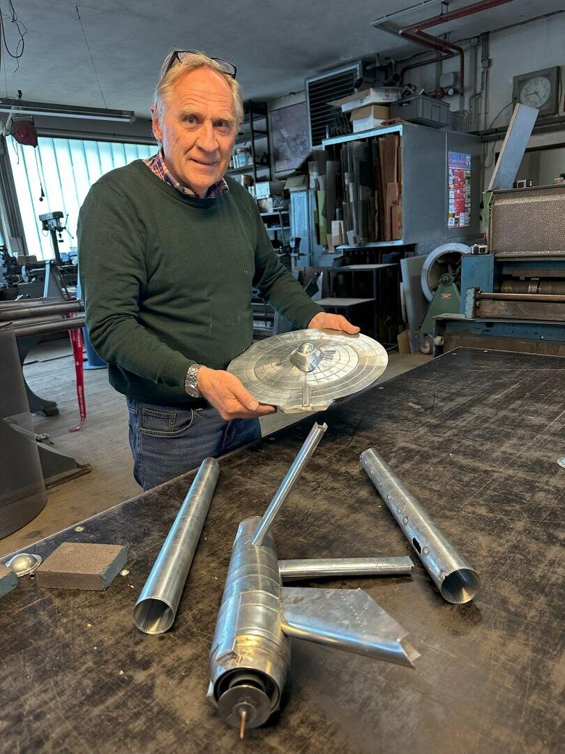 Ein älterer Mann hält in einer Werkstatt eine Metallscheibe, während er an einem Tisch mit Metallteilen in Form einer Rakete und Stäben steht, umgeben von verschiedenen Werkzeugen und Maschinen.