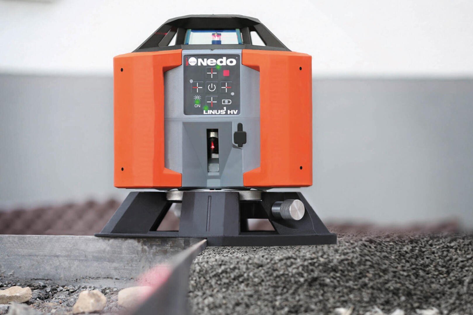 Eine Nedo LINUS HV Laser-Wasserwaage sitzt auf einem Stativ über Kies und wird zum präzisen Nivellieren und Ausrichten auf dem Bau eingesetzt. Das Gerät verfügt über ein digitales Bedienfeld und ein robustes orange-schwarzes Gehäuse.