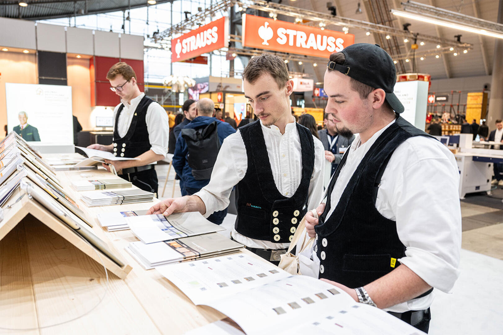 Three young men in traditional work vests check brochures at an exhibition table in a busy exhibition hall, Strauss signs can be seen in the background.