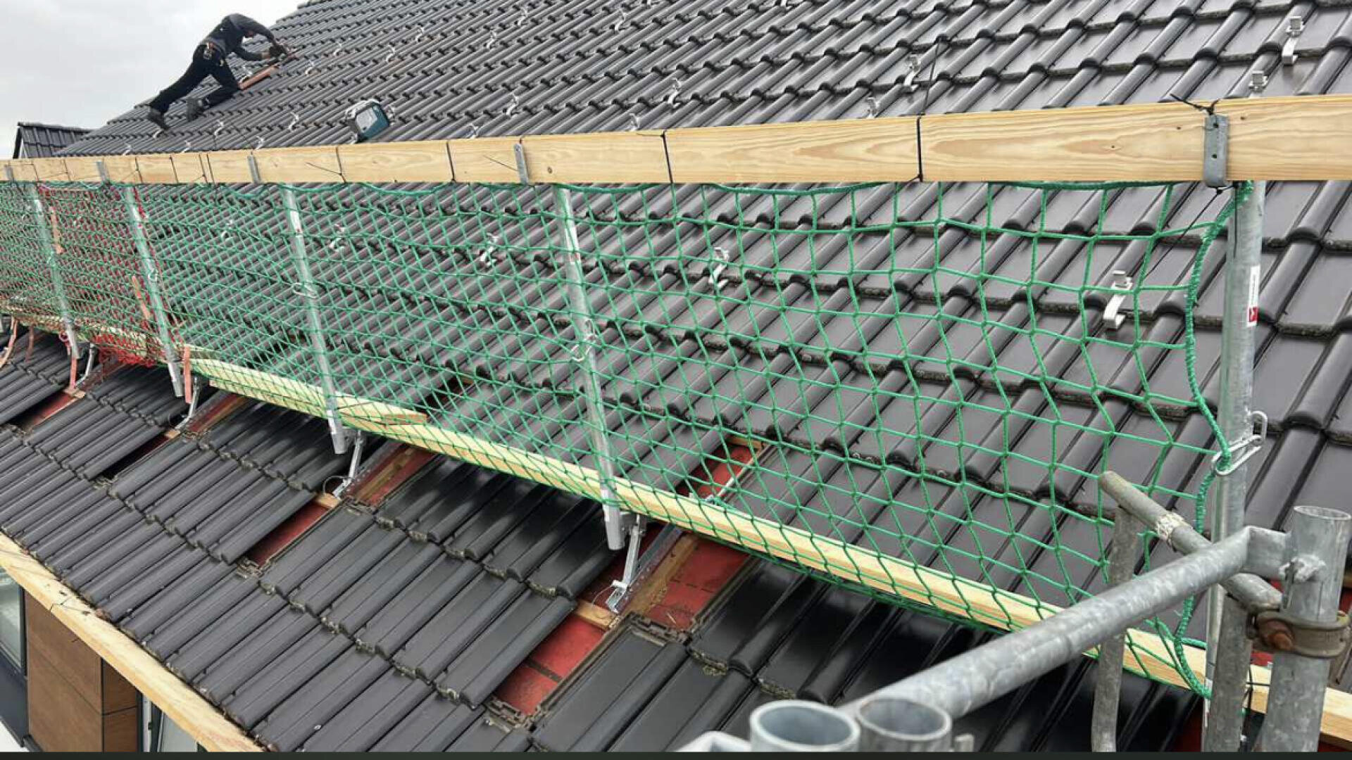 A worker on a sloped, dark-tiled roof installs or inspects safety netting and wooden boards attached along the edge, with scaffolding visible in the foreground.