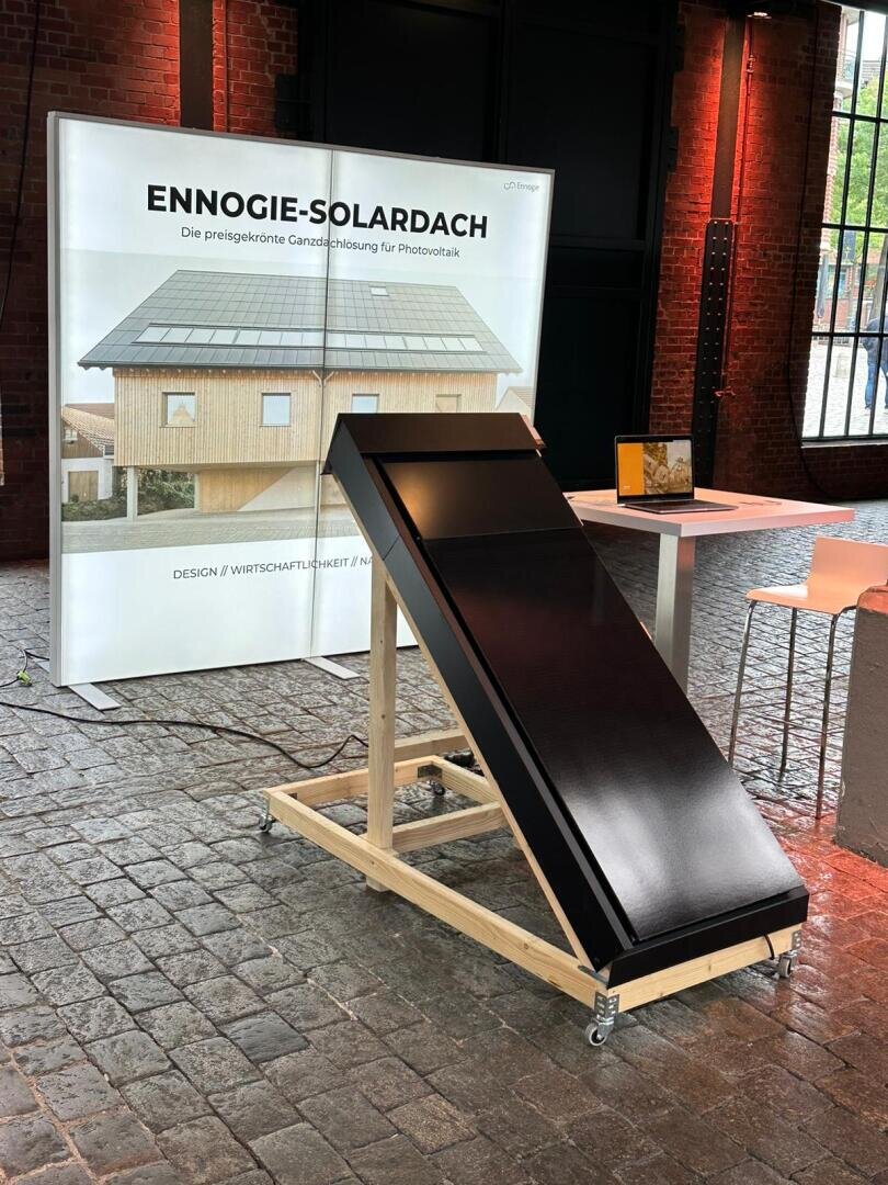 Ein elegantes schwarzes Solardachpaneel steht auf einem Holzständer in einem Raum vor einem Plakat, das ein Haus mit Solarzellen und die Aufschrift "ENNEOGIE-SOLARDACH" auf Deutsch zeigt. Der Raum hat Backsteinwände und große Fenster.