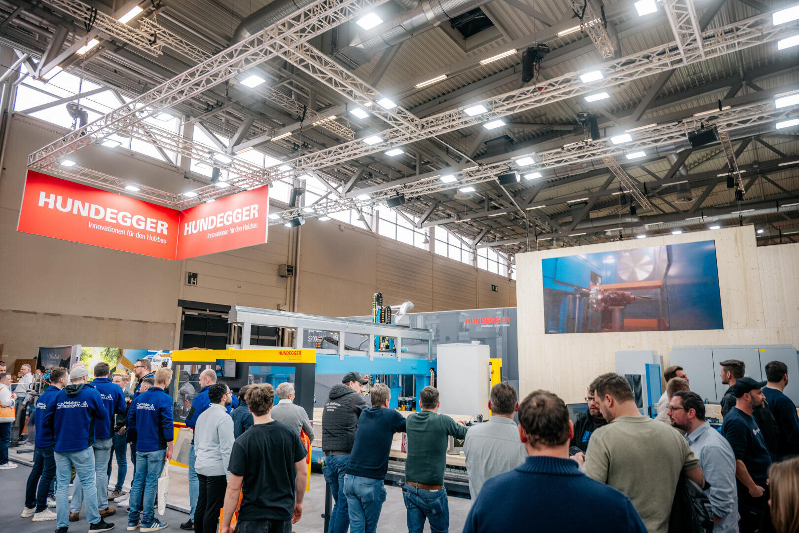 Eine Menschenmenge versammelt sich auf einer überdachten Industrieausstellung und betrachtet die Maschinen an einem Stand mit roten Hundegger-Schildern und einem großen Bildschirm, auf dem Geräte in Betrieb zu sehen sind. Helle Lichter erhellen den weitläufigen Veranstaltungsort.