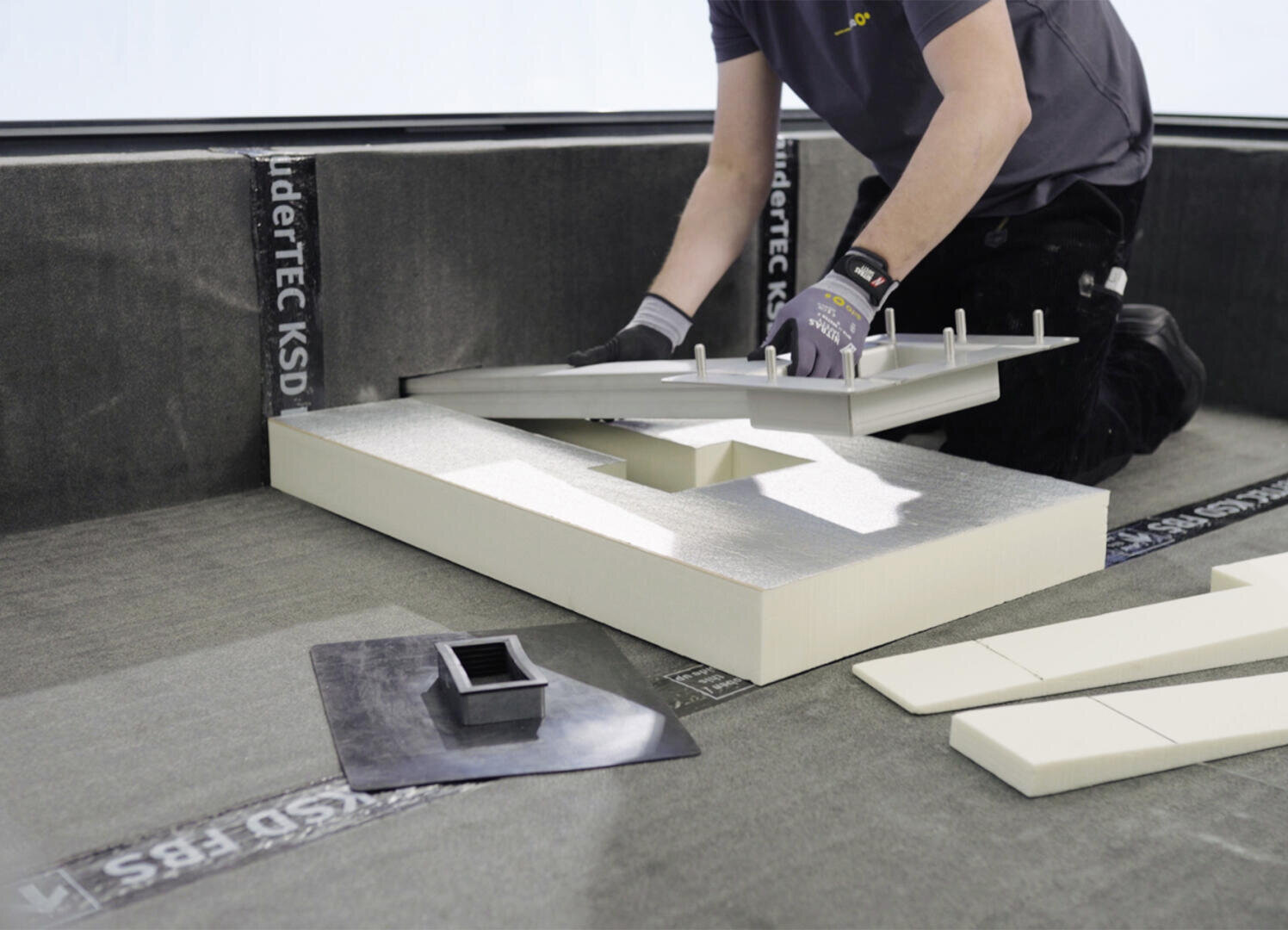 Eine Person, die Handschuhe trägt, verlegt auf einer ebenen Fläche eine Dämmung und ein Entwässerungselement und arbeitet mit Schaumstoffplatten und Baumaterialien.