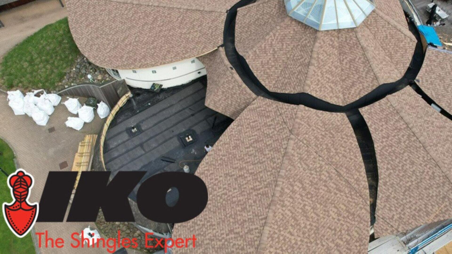 Luftaufnahme eines kreisrunden Gebäudes, auf dem gerade ein Schindeldach gedeckt wird; Bündel von Dachdeckungsmaterial und Ausrüstung sind zu sehen. Das IKO-Logo und der Text The Shingles Expert sind über das Bild gelegt.