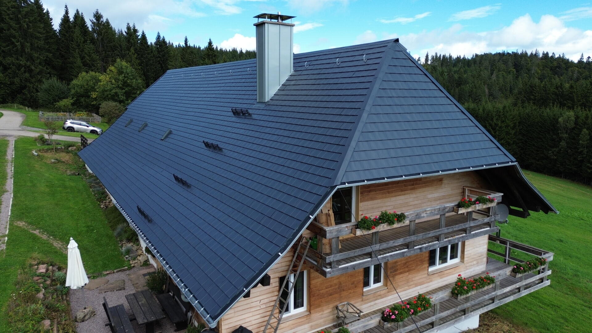 Ein großes Holzhaus mit einem steilen, dunklen Metalldach steht in einer grünen ländlichen Gegend, umgeben von Bäumen. Die Balkone sind mit Blumenkästen versehen, und ein weißer Sonnenschirm steht neben den Picknicktischen im Hof.