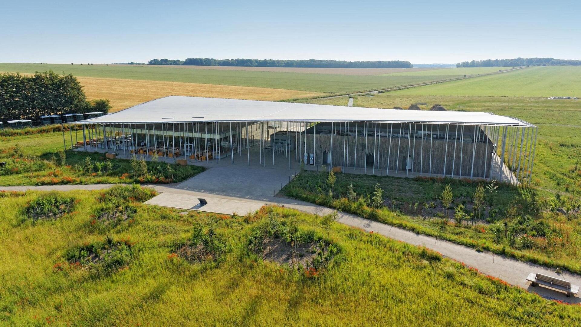 Ein modernes Gebäude mit einem großen, schrägen Dach und Glaswänden steht in einer grasbewachsenen, ländlichen Landschaft mit Feldern und Bäumen im Hintergrund. Gepflasterte Wege führen zu seinem Eingang.