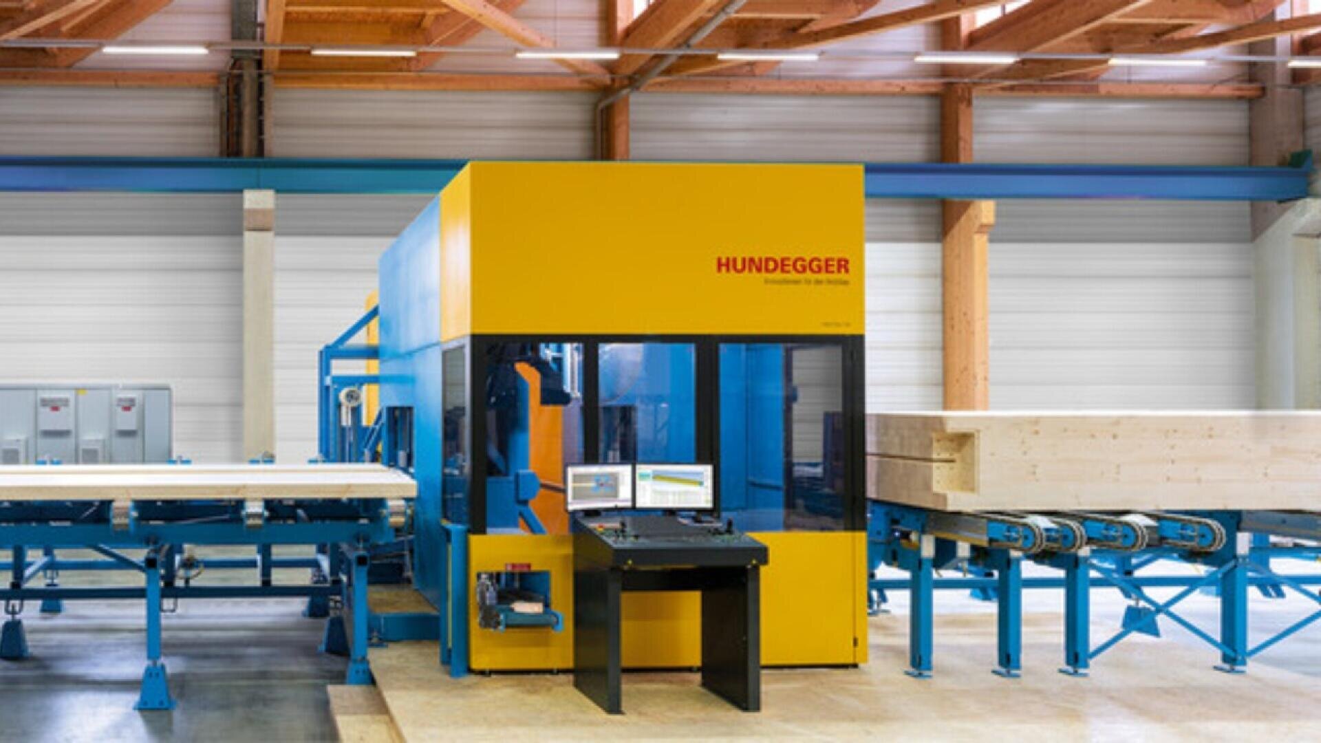 A large yellow and blue Hundegger industrial woodworking machine is positioned in a factory with wooden beams and conveyor belts transporting stacked lumber through the equipment. A computer workstation is set up in front of the machine.