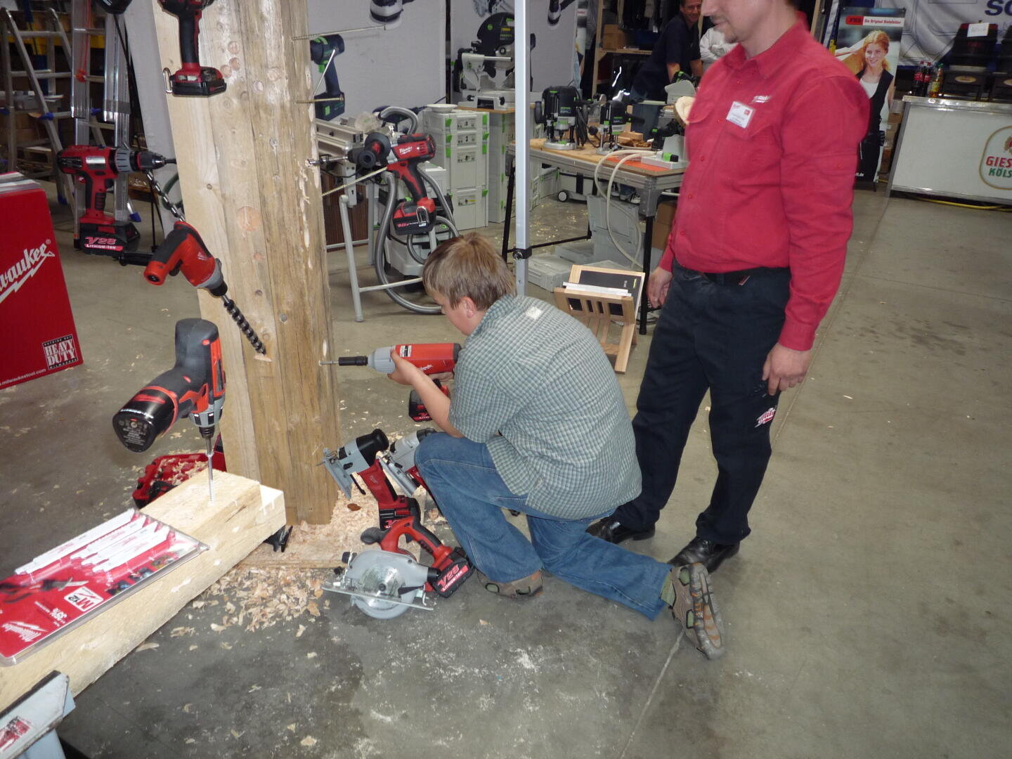Eine Person kniet auf dem Boden und bohrt mit einem roten Elektrowerkzeug in einen großen Holzbalken, während eine andere Person in einem roten Hemd in der Nähe in einer Werkstatt steht.