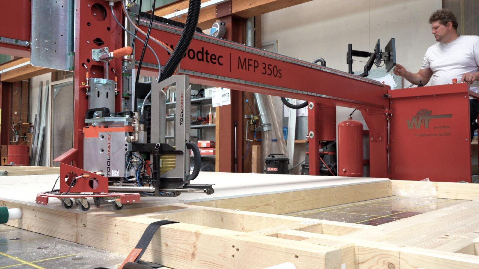 A man operates a large red wood processing machine labeled “MFP 350s” in a workshop, assembling or cutting wooden beams laid out in frames on the floor. Various tools and equipment are visible in the background.