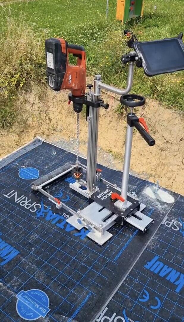 Eine rote tragbare Bohrmaschine, die auf einem vertikalen Metallständer mit einer Führungsschiene montiert ist und im Freien auf einer schwarzen Schutzfolie steht. Ein Tablet oder ein Bildschirm ist mit einer Klemme zur Überwachung oder Steuerung am Ständer befestigt.