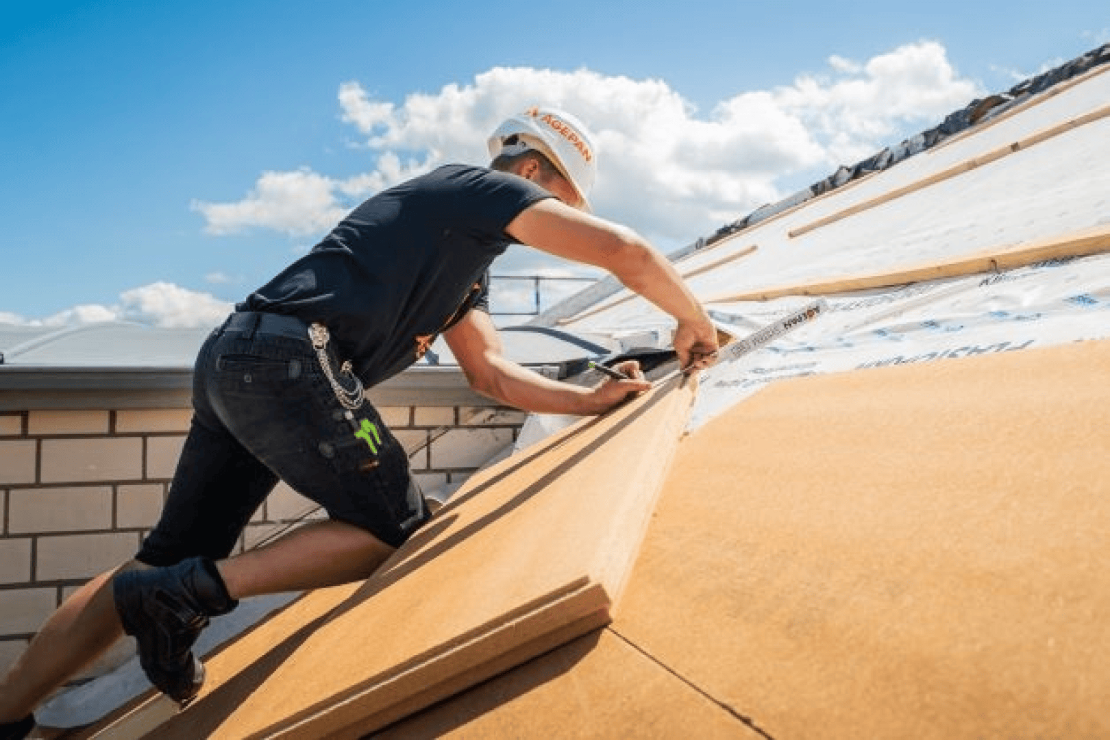 Ein Bauarbeiter, der einen Schutzhelm und kurze Hosen trägt, montiert bei strahlendem Sonnenschein Dämmplatten auf einem Schrägdach.