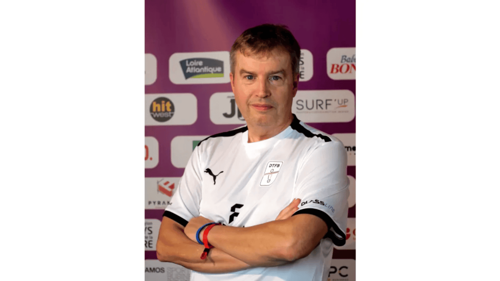 A man in a white sports jersey with black details stands with arms crossed in front of a backdrop with various sponsor logos. He has short brown hair and wears wristbands on both wrists.