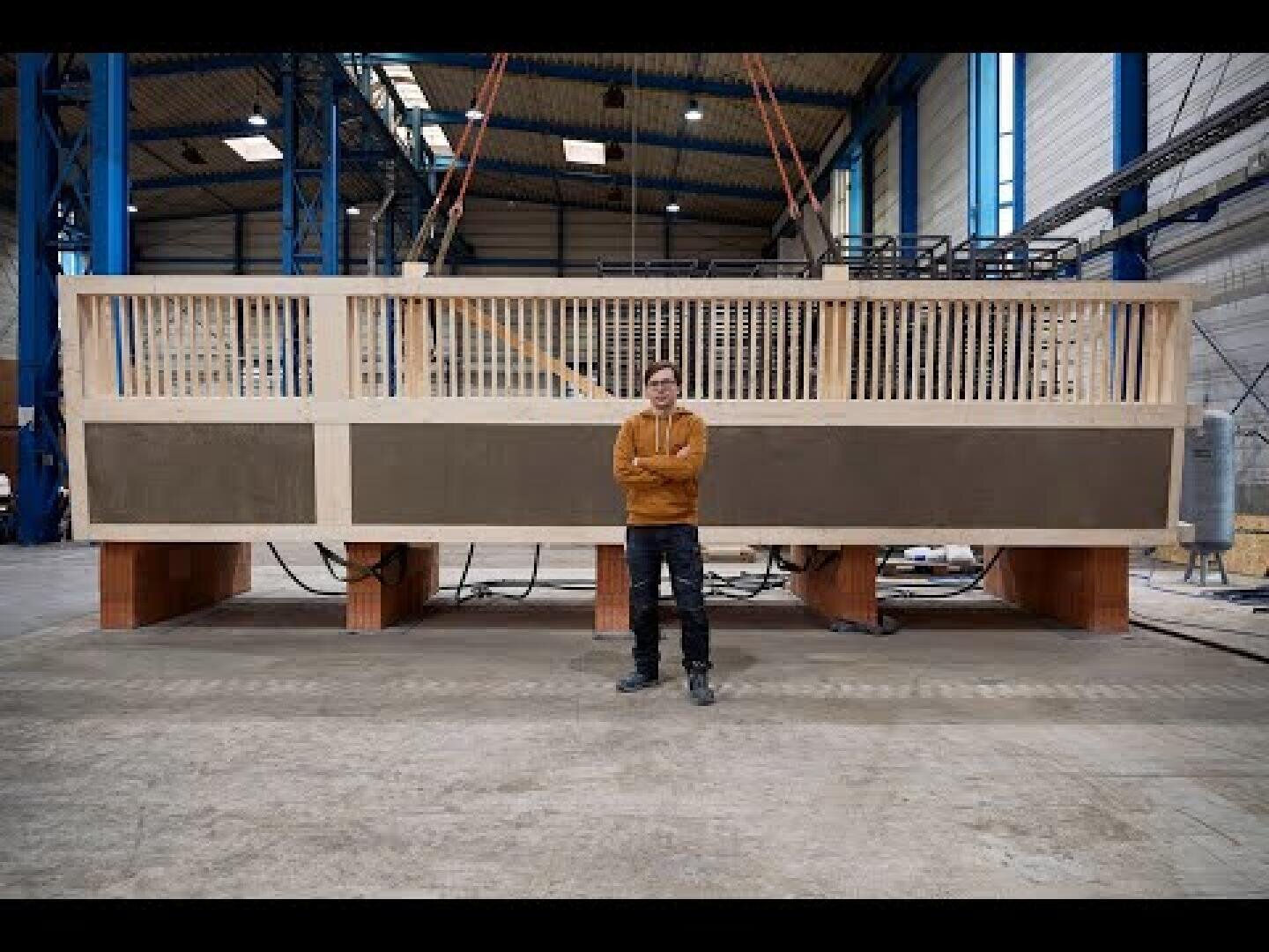 Eine Person steht mit verschränkten Armen vor einer großen, rechteckigen Holzkonstruktion in einer geräumigen Industriehalle. Die Struktur besteht aus einem gitterartigen oberen Teil und massiven Platten darunter.