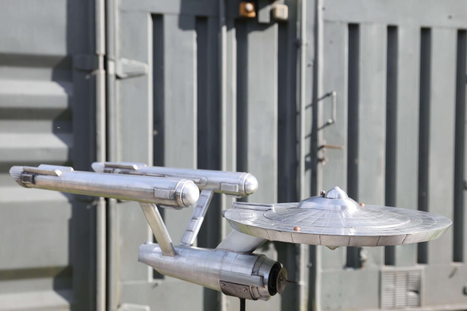 Ein silbernes Modell eines futuristischen Raumschiffs, das der Star Trek USS Enterprise ähnelt, ist im Freien vor einer grauen Metallwand angebracht.