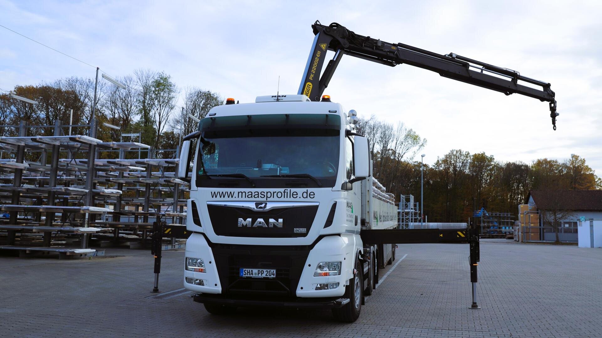 Ein weißer MAN-Lkw mit einem montierten Kran steht auf einem Industriegelände, umgeben von Metallgestellen und Bäumen im Hintergrund. Der Kranarm ist ausgefahren, und die Website maasprofile.de ist auf dem Lkw zu sehen.