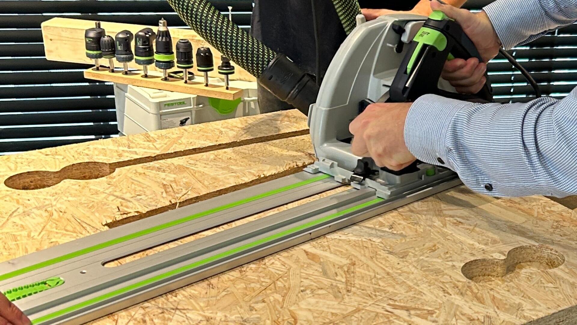 Eine Person schneidet mit einer Raupenkreissäge eine OSB-Platte auf einer Werkbank zu, während im Hintergrund Bohrer und Werkzeuge zu sehen sind.