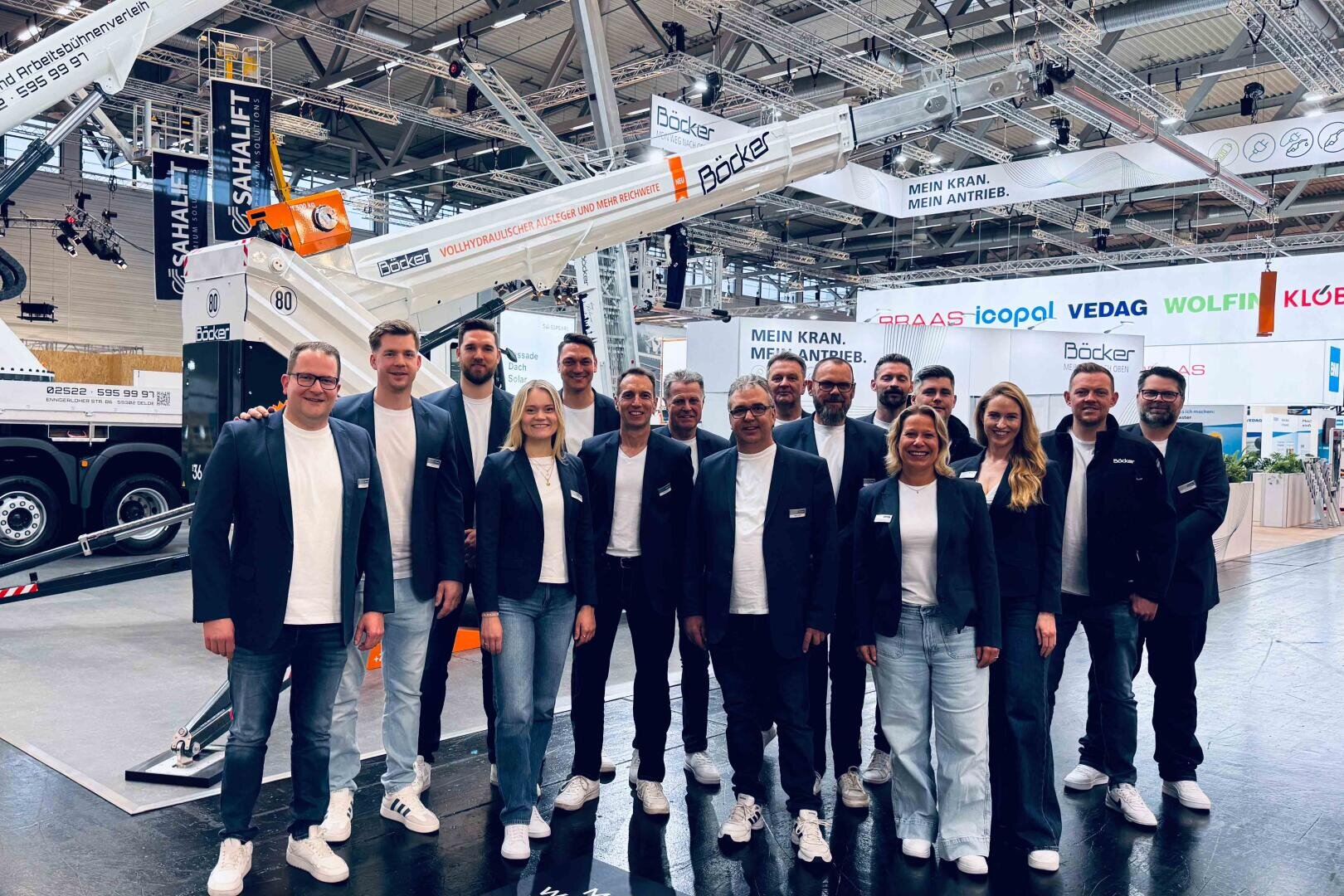 Eine Gruppe von dreizehn Personen in legerer Business-Kleidung und mit Namensschildern posiert zusammen und lächelt vor einem großen Kran auf einem Messestand in einer Halle.