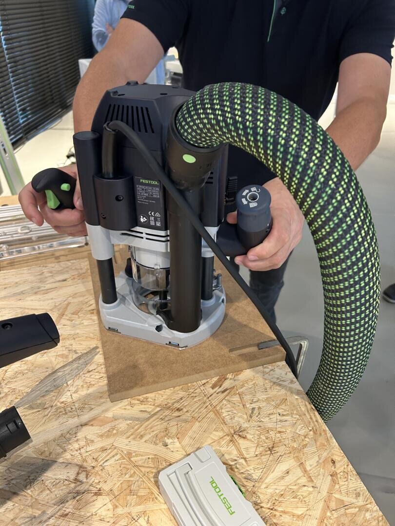 Eine Person benutzt eine handgeführte elektrische Oberfräse mit einem grün-schwarzen Schlauchaufsatz, um ein Stück Holz auf einer Werkbank zu bearbeiten. Werkzeuge und Zubehör sind in der Nähe zu sehen.