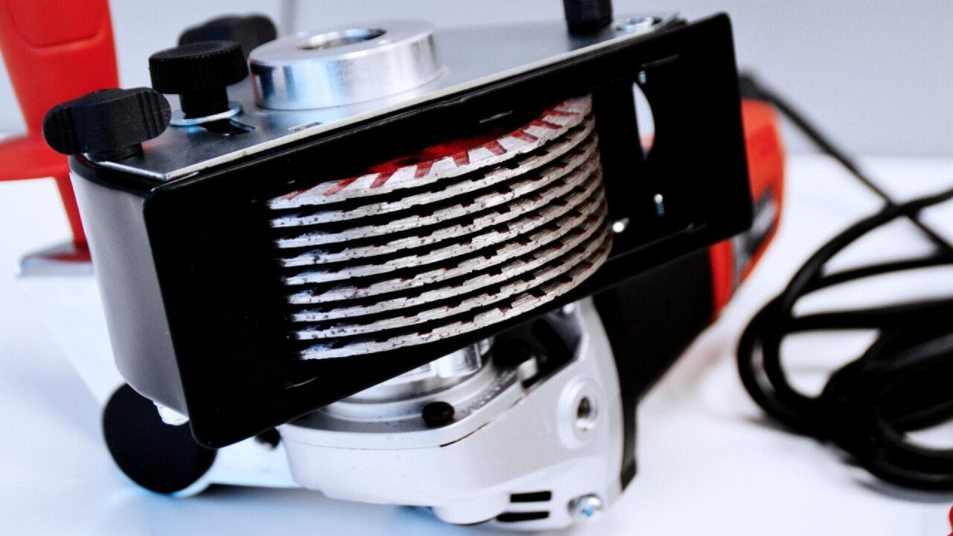 Close-up of a power tool featuring multiple stacked circular saw blades, a red handle, and a black cord, placed on a white surface.